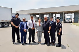 The Secretary and officials are standing in front of a bridge entry way. Trucks can be seen in the background. 